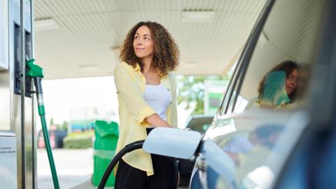 A stock image of a woman filling up her car with petrol. She has shoulder length hair and is wearing yellow shirt, white t-shirt and black trousers. The car is blue. 