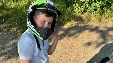 Oliver sat on his bike wearing a green and white cycle helmet and looking towards the camera. There are trees and shrubs in the background. 