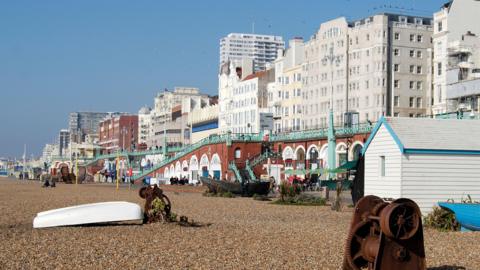 The image shows a boardwalk and beach with cafes and restaurants on seafront; an overturned white boat can also be seen on the rocks.