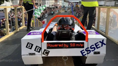 The back of a cart with BBC Essex branding written across it. Crowds are lining the street.