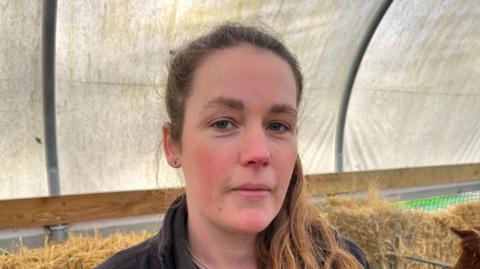 A head and shoulders image of Kate Ockleton. She is wearing a dark grey jacket over a dark coloured top and standing in front of some hay. She is looking directly into the camera.