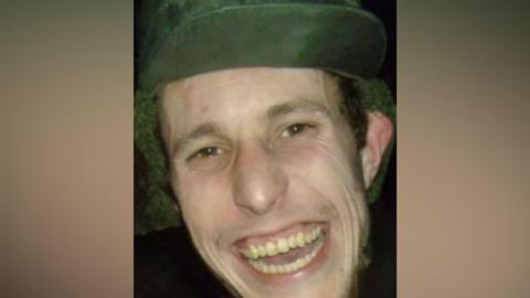 A man wearing a green cap is pictured grinning at the camera. He has brown eyes and some brown stubble around his cheeks and chin.