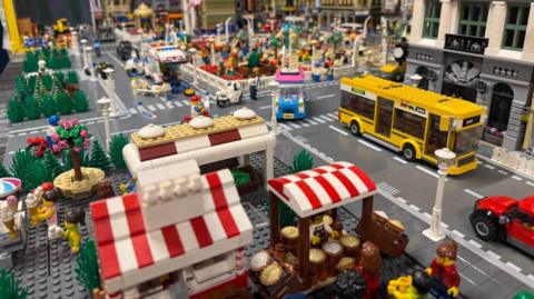 A large Lego town display, with a yellow bus, market stalls, cars, crossings, buildings and people, all on a grid system.