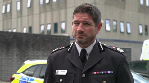 Jim Colwell is standing outside of a police building which a police vehicle in the background. He has dark hair and is dressed in uniform. 