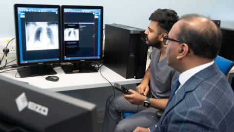 Two men are looking at a screen on the left. The screen has pictures of chest x-rays. One man is wearing hospital scrubs while the other is wearing a grey suit, white shirt and blue tie. 