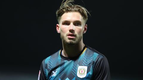 Joel Tabiner is wearing his back and light blue Crewe shirt with the club's badge in white, yellow and red on the left-hand side of his chest and is looking into the distance during a night game. The background is completely black.