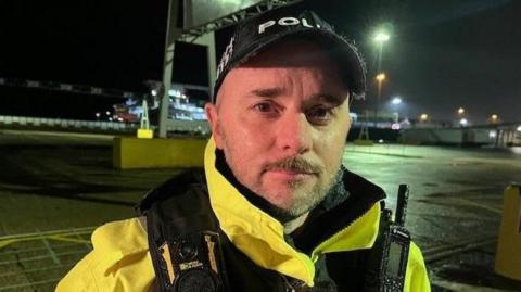 Sgt Ross Haybourne is wearing a police hi-vis jacket and black police cap. He is standing in a dark concreted area with port machinery and buildings in the background. 