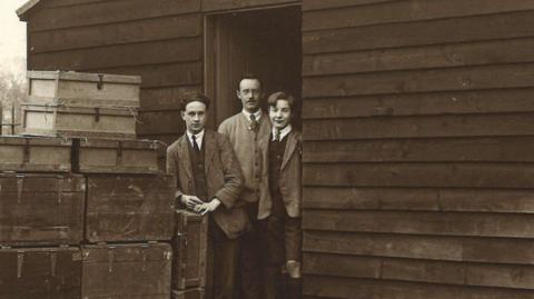 An old black & white picture showing the doorway of a large wooden hut with three men stood in it, dressed smartly in jackets and ties. There are a stack of trunks and boxes outside, presumably filled with books