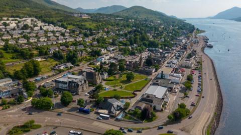 An aerial view of Fort William and Loch Linnhe on a sunny day in Lochaber. There are houses on a hillside among trees, and down towards the waterfront larger buildings including churches and the A82 road.