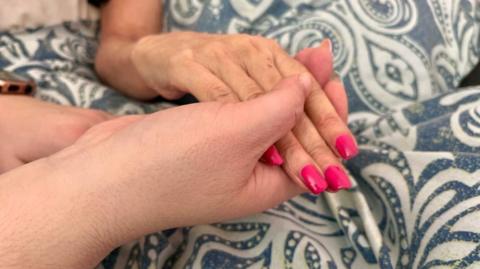 Two people are holding hands over a duvet cover. One hand is of an older woman with pink painted nails. The other hand is also a woman's.