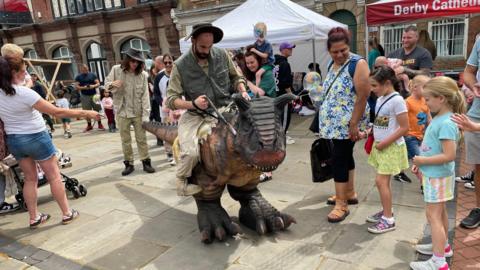 A man riding a plastic dinosaur while young children look on