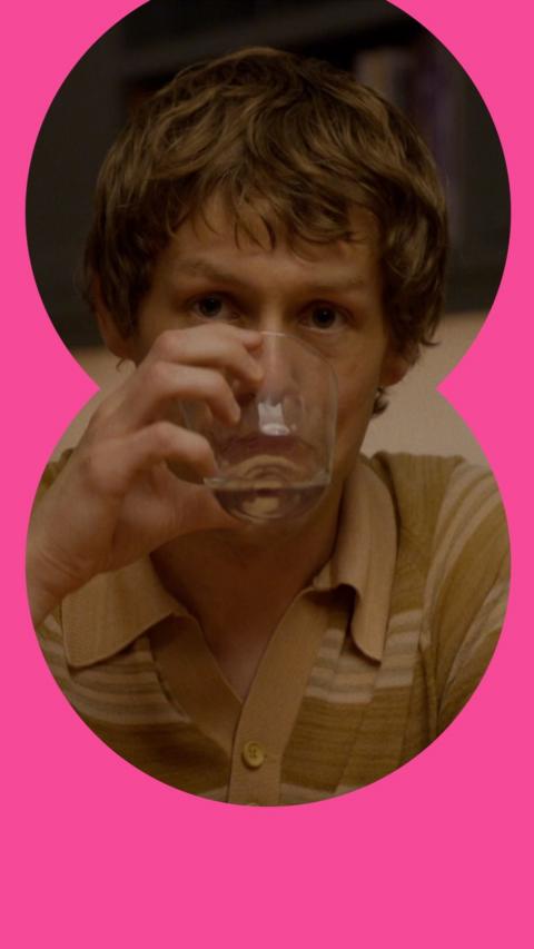 A man drinks from a glass at a dinner table. He has short brown hair and wears a brown patterned T-shirt