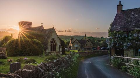 A picture of the centre of the Cotswold village of Snowshill at sunset. The sun can be seen going down with part of it obscured by the village church. A road bordered by stone walls runs between the church and its graveyard and houses opposite. There is extensive foliage with purple and pink flowers on the houses and wall
