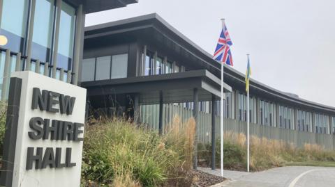 A council building with a large square stone sign outside saying "New Shire Hall" in dark grey lettering against a beige background. Two flagpoles outside fly the British flag and a yellow and blue one.  There are decorative grasses outside the building which is modern with glass and vertical shading.
