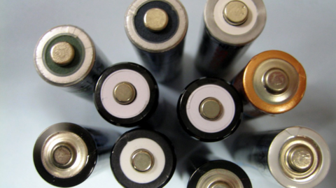 The image shows a close-up bird's-eye view of several cylindrical household batteries close together. Each battery is positioned upright so the positive terminal faces the camera. The terminals appear as small, raised metal circles surrounded by different coloured rings, including white, black, green and gold. 