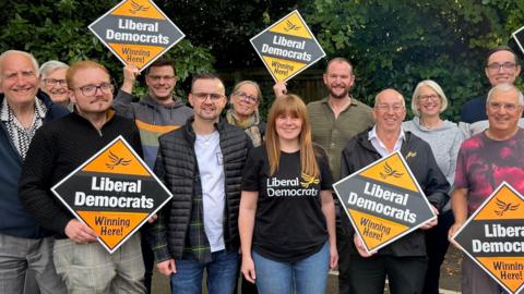 A group of Liberal Democrat candidates and existing councillors. The picture shows nine men and three women. Five of them are holding up boards which read "Liberal Democrats Winning Here!"