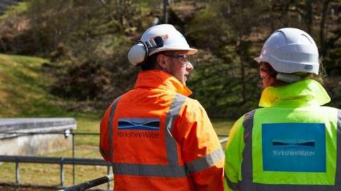 A man and a woman wearing hard hats and high-visibility jackets with the Yorkshire Water logo look over a sewage plant.