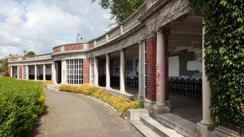 Sun Shelter, Cliff Gardens, Westcliff Parade, Westcliff-on-Sea. The single-storey  shelter is curving away and has glazed panels, columns and balustrade above. In front of it is a pavement. 