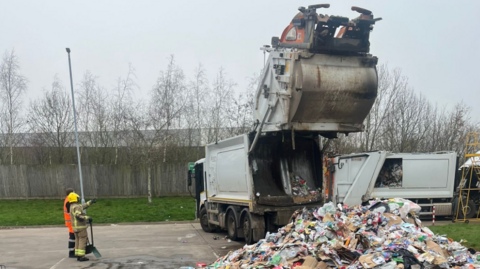 A pile of rubbish can be seen in front of a fire damaged bin lorry. A firefighter and another person are looking at the lorry which is parked in the open with grass surrounding it.
