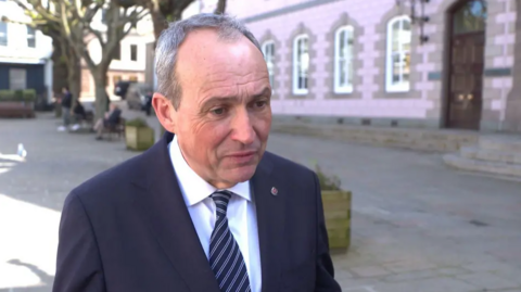 A man wearing a black suit with a striped navy and grey tie standing in the square outside of a government building.