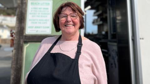 Rhona Hewit is smiling while standing in front of her kiosk. She is wearing a black apron and pink top.