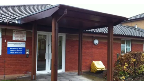 The entrance of a one-storey, redbrick building with white PVC double doors. There are four pillars holding up a roof coming out from the doorway. A blue and white sign with the NHS logo on it reads 'Broadway Park, Wessex House, Main Entrance'.