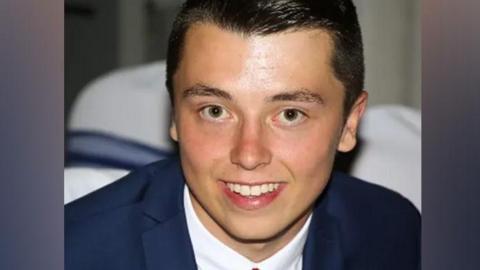 Connor Brown looking directly into the camera and smiling. He has dark hair, brushed to his right, and is wearing a navy suit, white shirt and red tie.
