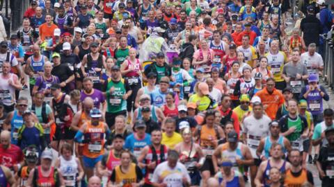 Hundreds of runners seen competing in a marathon. The people running are a mixture of ages and genders. They are wearing different coloured running tops.