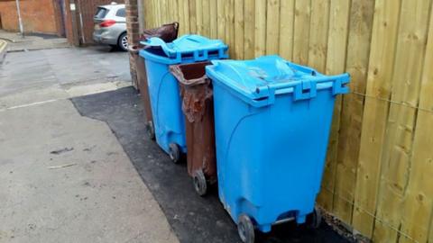 Four brown and blue wheelie bins against a wooden fence showing signs of fire damage with the lids of all four melted 