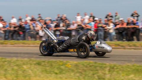 A man wearing black leathers and a helmet lying down on a three-wheeled vehicle that is travelling at speed. There is a blurred crowd watching on from behind metal barriers.