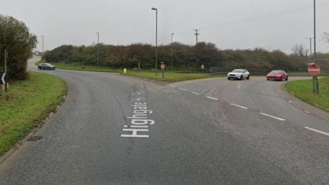 Google Street View image of Highgate Hill roundabout in Indian Queens, Cornwall. A white car and a red car are approaching the give way lines at an exit which also has two no entry road signs either side of the road. A grey car is coming round the roundabout towards the exit, but looks like it might be about to exit the roundabout. It is a grey cloudy day.