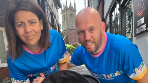 Mark and Natalie McLeod in blue running tshirts, they are standing next to a building with a sign that says tattoo collective, and behind them is a church tower