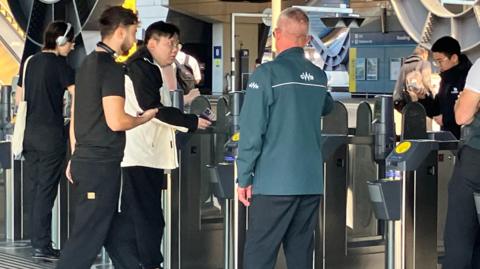 Confused looking commuters around ticket barriers at Reading station. A man in a GWR jacket offers advice. 