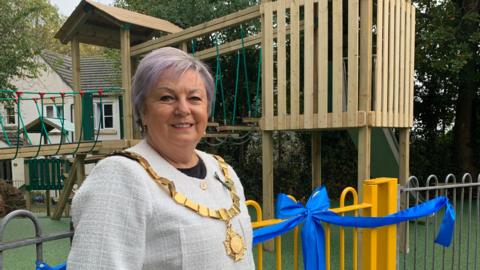 Mayor of Saltash Councillor Julia Peggs pictured in front of Grassmere Way play areas. There is a blue ribbon on the gate and a wooden play set in the background.