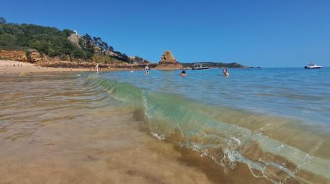 A few people are swimming in a blue sea at Les Creux. There are several small boats off the coast and the sky is blue and cloudless. In the distance several people can be seen relaxing on the sandy beach.