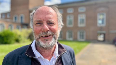 Liberal Democrat leader Julian Brazil standing in front of County Hall and wearing a white shirt, a maroon zip-through jumper and a navy blue blazer