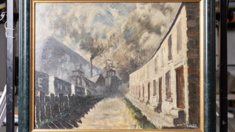 Colliery Row by Norman Cornish in a black frame with a golden edge. The paining portrays a row of terraces houses on one side, with minework buildings opposite, with smoke coming out of a large industrial chimney and the smoke filling the sky.