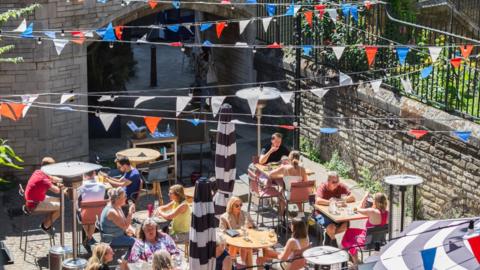 Diners eat in a sunny restaurant garden. Red, white and blue bunting is hanging above them.
