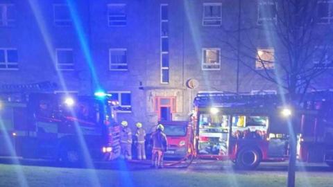 Firefighters next to fire engines in darkness with their blue lights shining.