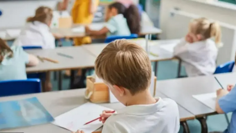 A young boy writes on a piece of paper in a classroom. There are other children sat on desks nearby. The boy has short brown hair and a white top on with his back to the camera. Other pupils are also wearing white tops and are blurred in the background. 
