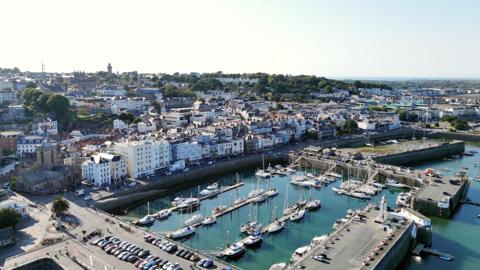 An aerial shot of St Peter Port in Guernsey. It shows boats in the harbour and a road with parked cars. Buildings can be seen in the background. 
