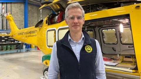 Charles Hackett wearing a navy Devon and Somerset Air Ambulance branded gilet, a blue and white gingham checked shirt and clear framed glasses, with his grey hair styled slightly in a a short quiff. He is standing in front of a yellow air ambulance, which has its door open and there are some books and tools on the floor of it. It is in a warehouse/hangar with brick walls and a light coloured floor.