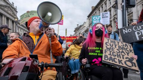 People in wheelchairs are pictured at a demonstration in central London with various signs, the most prominent of which reads 'they say cut back we say fight back' and 'welfare not warfare', and one of the protesters is holding up a megaphone. There are also other people standing behind them protesting as well and the sky is cloudy. Taken in London on 26 March.