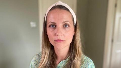 Julie Imrie looking straight ahead with a neutral expression. She has long, dark blonde hair and is wearing a white headband and a green shirt with blue on. 