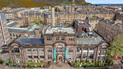 An aerial shot of the Summerhall arts complex in Edinburgh on a sunny day during the Festival Fringe with sections of the Old Town and part of Arthur's Seat in the background.