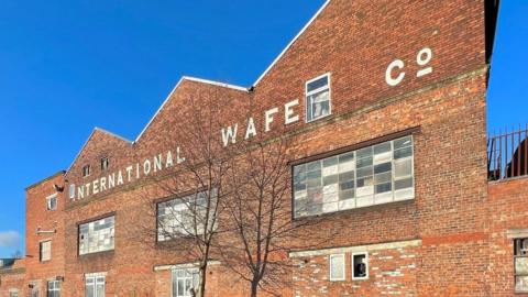 Progress Works, Ayres Road, Old Trafford, a red brick building with a tree in the foreground on a sunny day with the words 'international wafe co' emblazoned on the front of the building, 