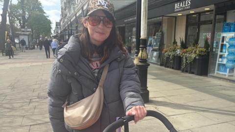 Megan Pennington, 33, who has a baseball cap with a NY logo, large framed dark red sunglasses, and a grey puffer coat, smiles at the camera while pushing the handle of a pram.