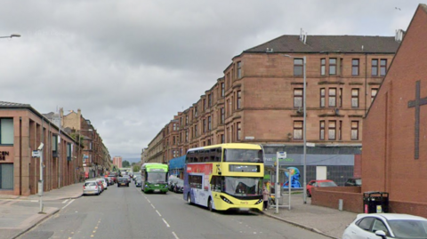 A city street, with red brick buildings and tenement flats on either side of the road. Cars and buses are driving up and down the road.