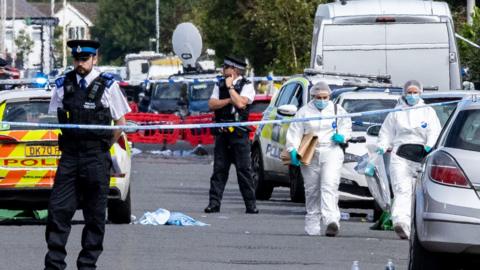 Police and forensic investigators in white protective clothing and masks are seen walking on a road filled with police vans and cars, with debris strewn across it and sealed off by blue and white police tape.