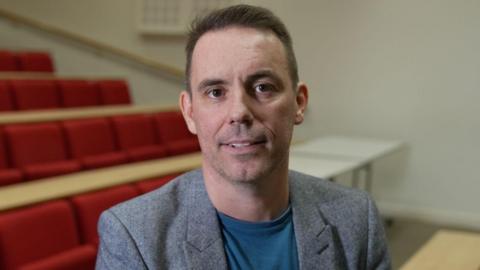 The principal of Dundee and Angus College Simon Hewitt. He has short black hair and brown eyes. He is wearing a grey suit jacket and light blue T-shirt 
